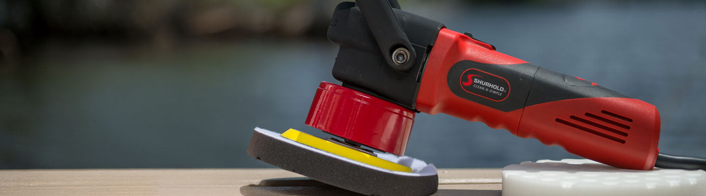 A red and black electric polisher with a foam pad attached rests on a wooden surface near water.