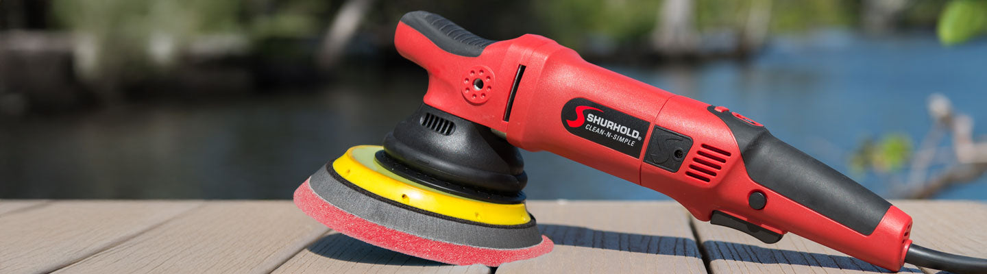 A red and black electric polisher with a yellow and black foam pad resting on a wooden surface outdoors near a body of water.