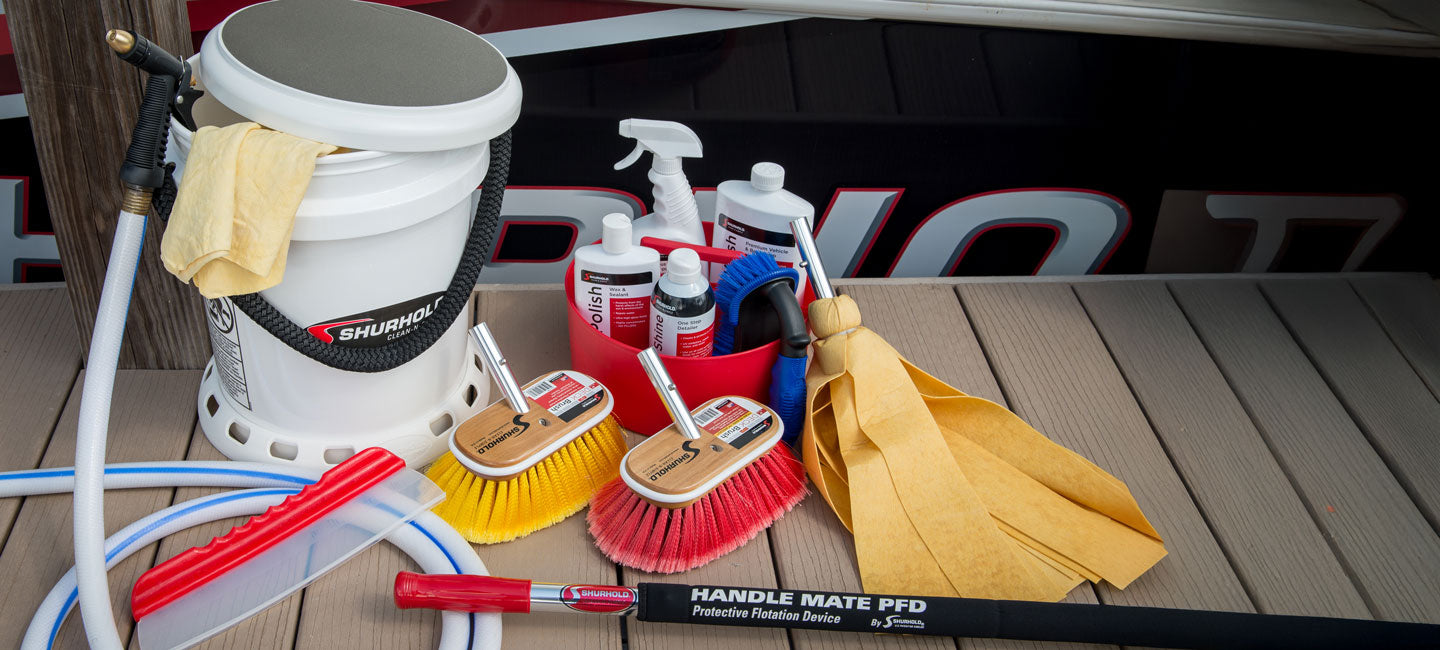 A variety of boat cleaning supplies including brushes, spray bottles, a bucket, towels, a scrubber, hose, and a handle are arranged on a wooden surface.