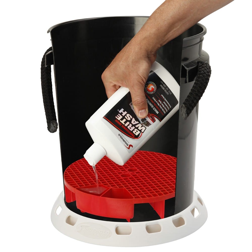 A hand pours car wash solution from a white bottle onto a Shurhold Industries, Inc. Bucket Grate with an integrated baffle system, placed inside a black bucket with rope handles.