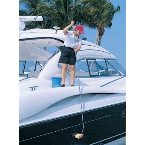 A person in a white shirt, black shorts, and red cap is standing on a boat by palm trees, cleaning the hull with a brush at a 30-degree angle using the Curved Adapter from Shurhold Industries, Inc.