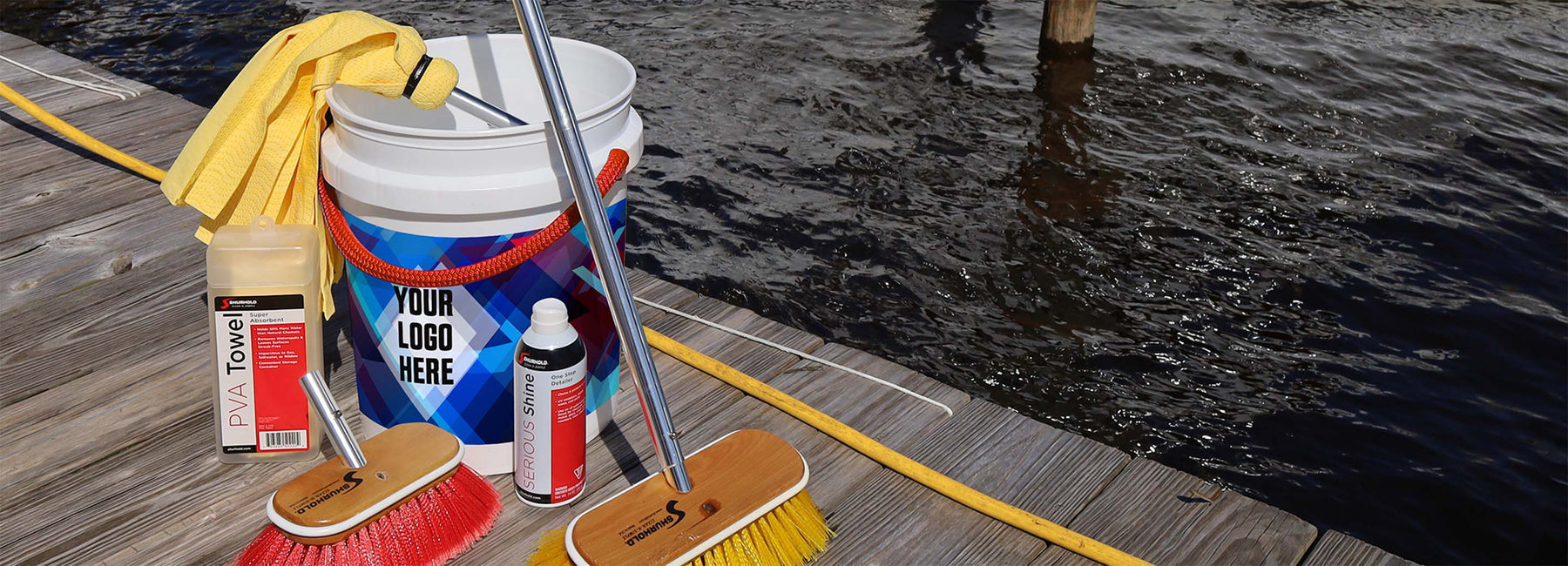 Cleaning supplies, including two brushes, a towel, a bucket with a blank logo space, a bottle, and a container, are arranged on a dock beside water.