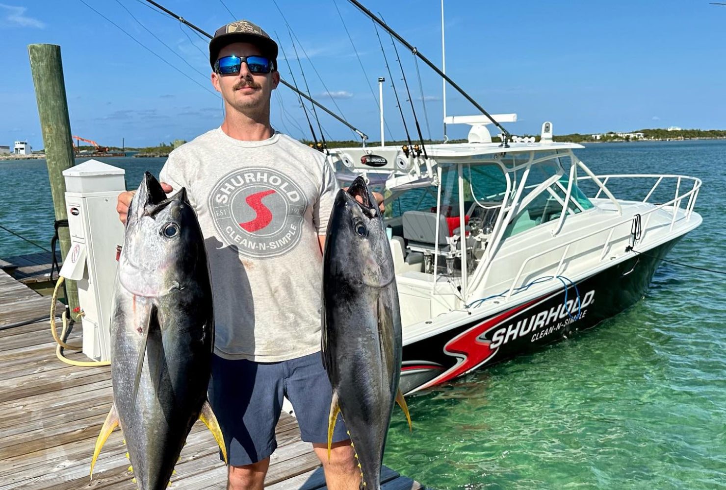 A man stands on a dock holding two large fish, with a fishing boat labeled Shurhold Clean-N-Simple in the background on clear water.